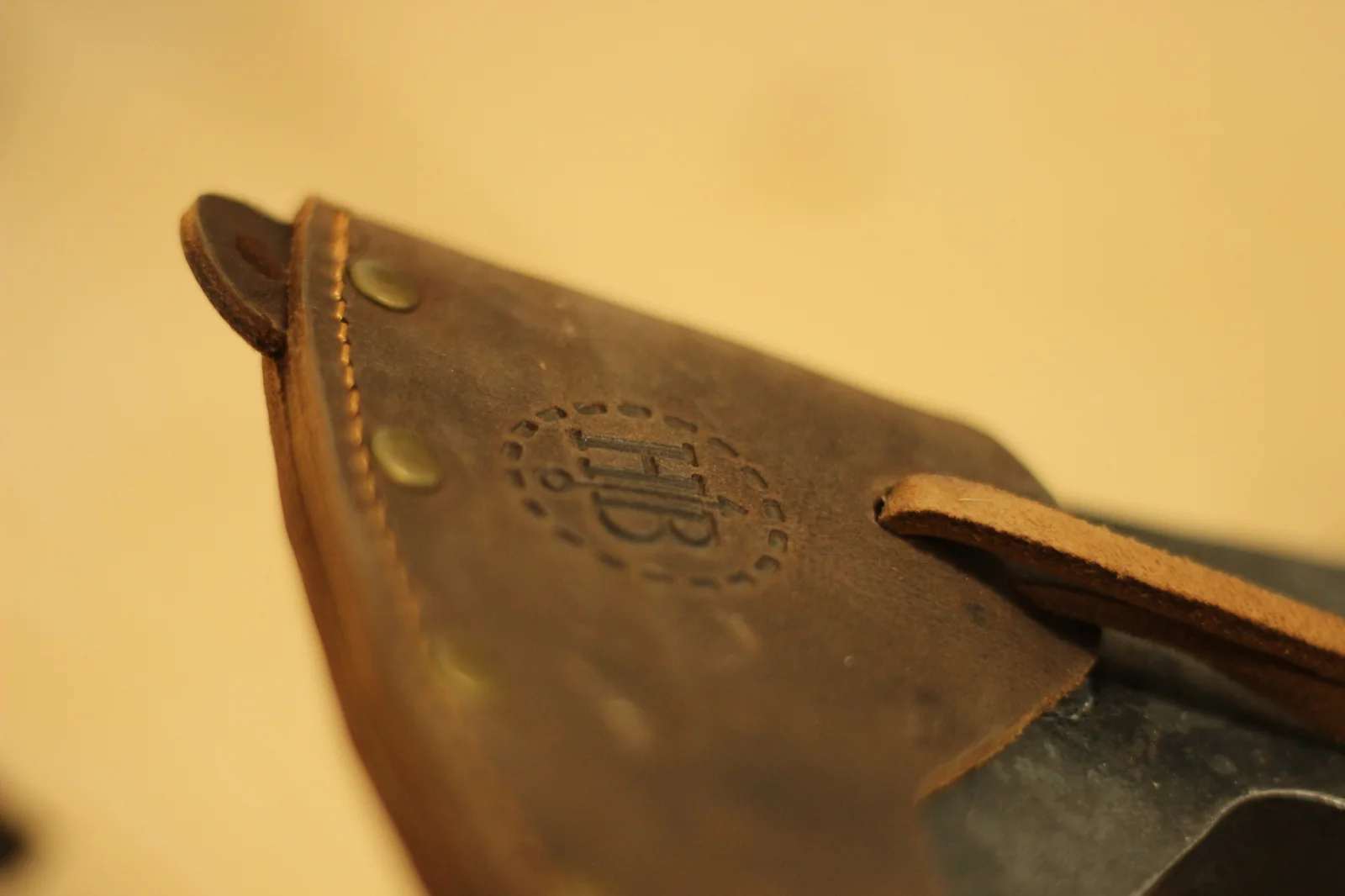 Macro shot of the HB hallmark embossed into the leather sheath surrounded by brass rivets and hand stitching