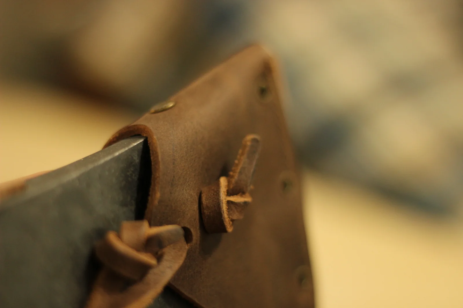 Close-up of the leather sheath at the cutting-edge corner showing the leather strap and brass studs that secure the strap to the sheath body