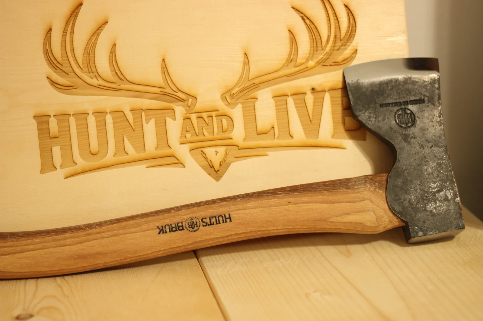 Hults Bruk Dvärdala axe leaning at an angle in front of the Hunt and Live laser-engraved wooden plaque with the deer skull and antlers