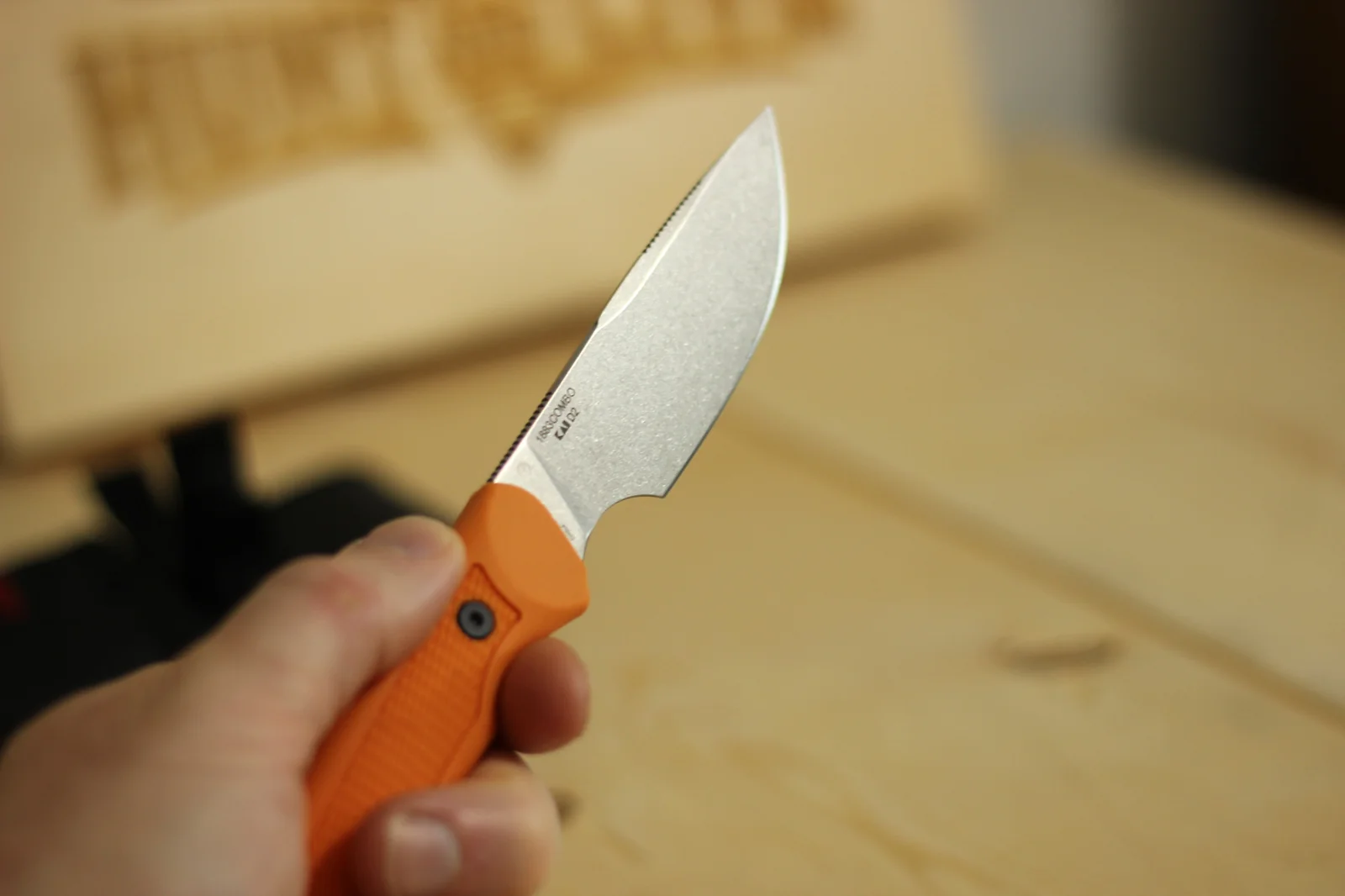 The 1883 knife held in hand showing the full drop-point blade profile with the Hunt and Live laser-engraved wooden plaque visible in the background