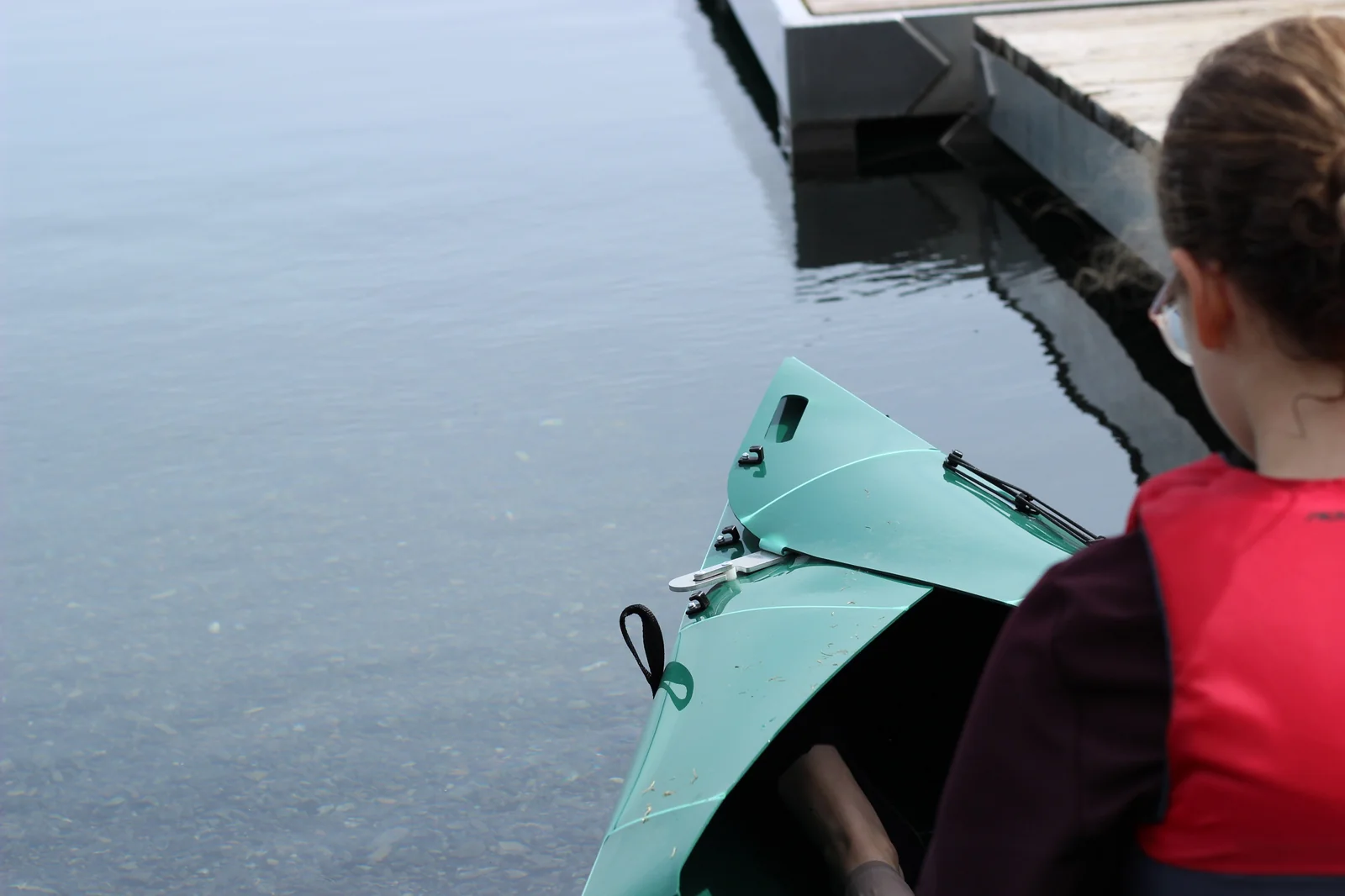 View from the dock looking down at the bow of the Tucktec Pro kayak in shallow water beside a woman in a red life jacket