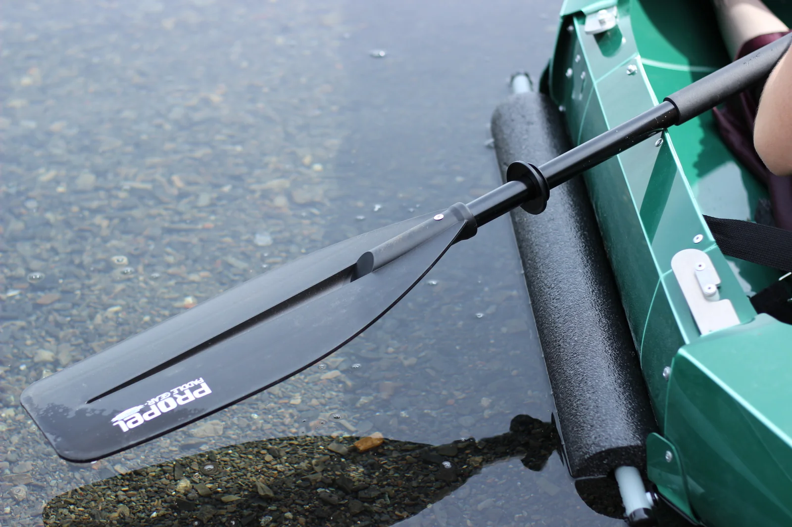 Close-up of the Tucktec 2-part paddle blade in clear shallow water beside the green hull and black foam stabilizer pontoon, with PROPEL branding visible on the blade