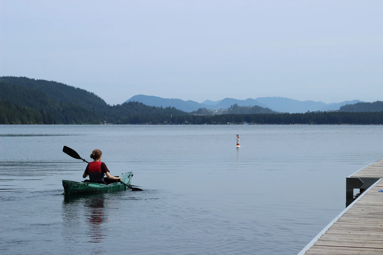 Paddler in a red life jacket paddling the Tucktec Pro kayak away from the dock toward a distant lakeshore with mountains and a navigation marker buoy visible